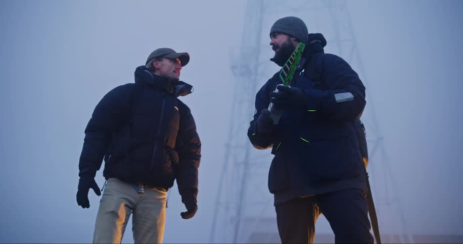 Two workers greet each other and have a conversation in an outdoor environment with an electrical transmission tower in the background at the end of their conversation they bid farewell and each goes in a different direction