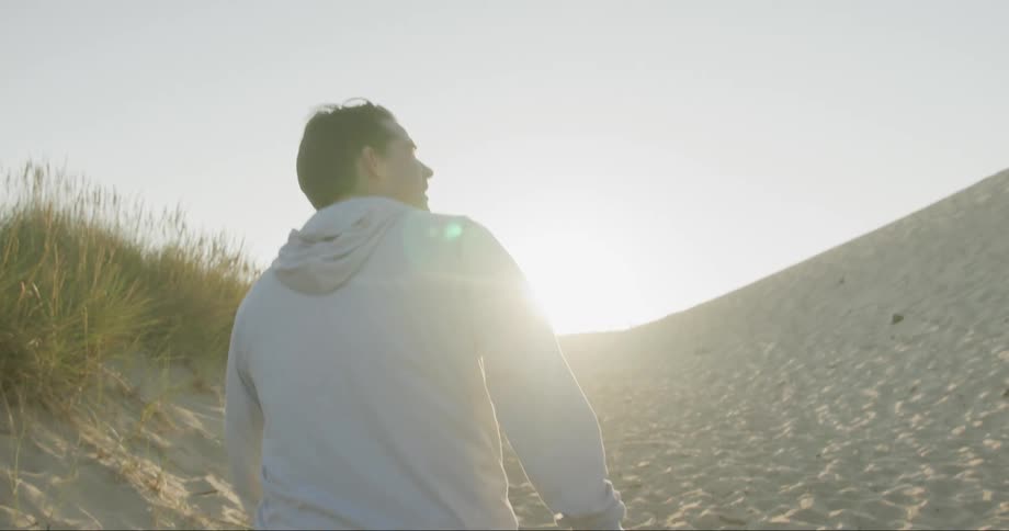 A man walks near a sand covered hillside with tall grass and weeds as the sun shines brightly in the distance