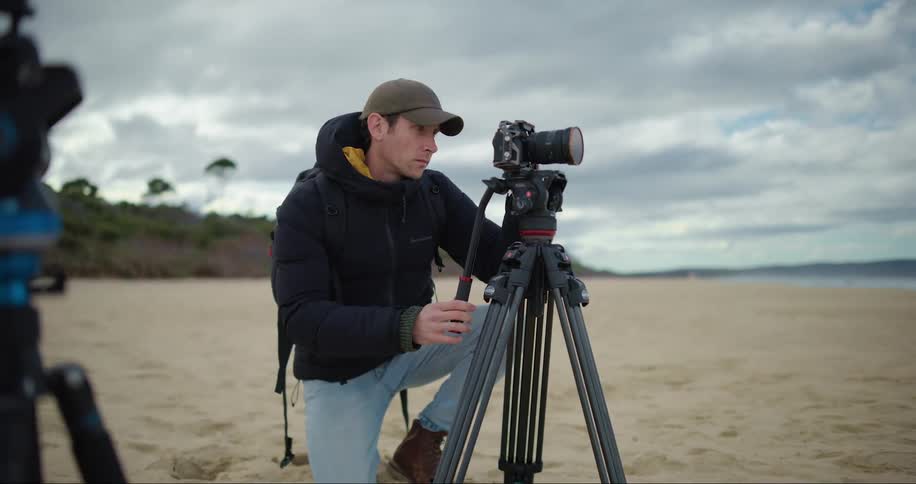 A male photographer setting up his camera and tripod on a beach during the day