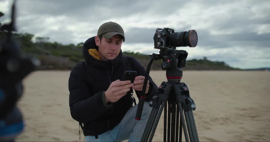 A photographer is on the beach kneeling near his camera and tripod as he takes his cell phone out od his pocket and looks at his camera as he puts the phone back in his pocket
