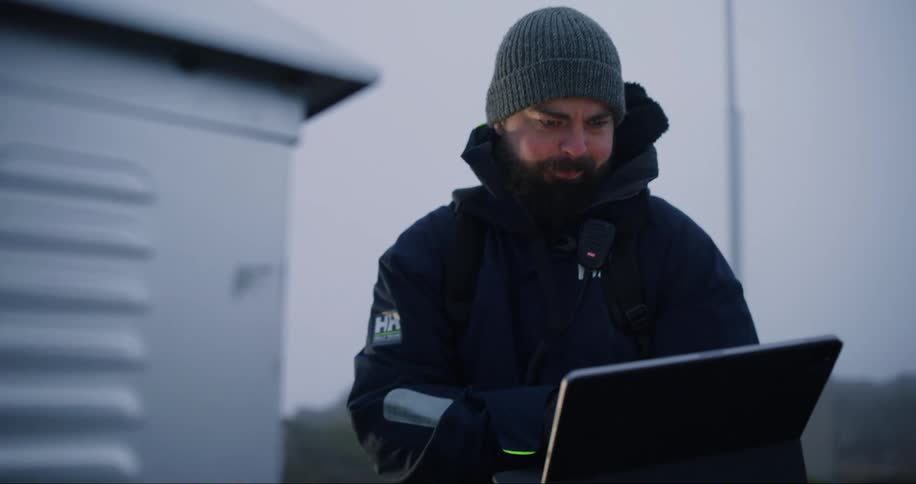 A man wearing a beanie and a heavy coat inspects equipment while holding a laptop