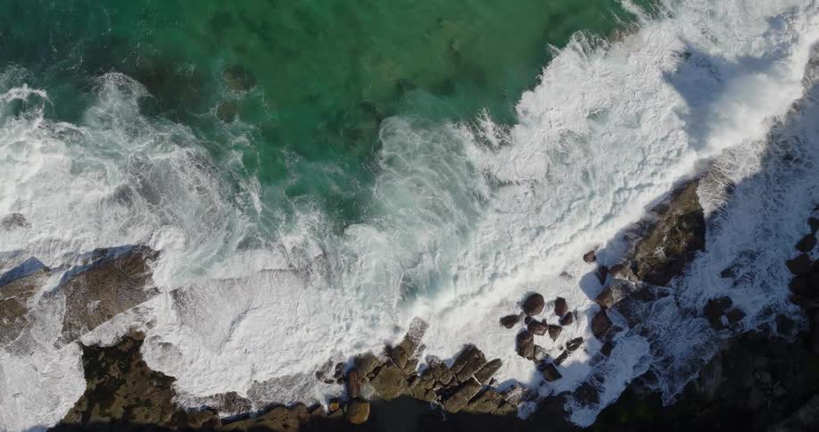 Drone view of ocean waves crashing against a rocky coastline during a bright sunny day