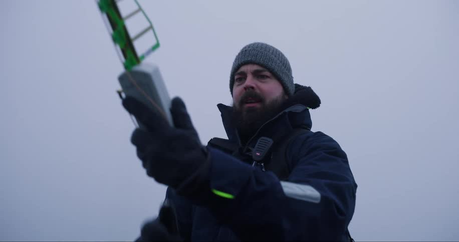 A bearded man in a cap and coat with a scientific instrument in his hand pointing upwards