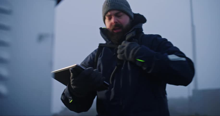 A man wearing a winter coat and hat holds a tablet computer as he looks at a utility box and speaks into a walkie talkie UHF radio on his coat and walks away