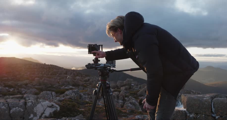 A man is standing on rock covered ground as he looks through a camera on a tripod and the sun shines over the mountains in the distance under a cloudy sky