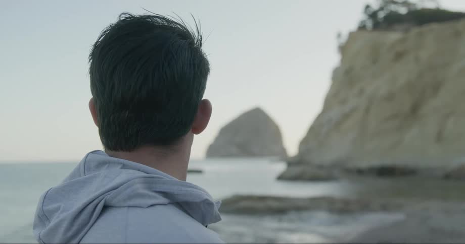 Man stands on coast looks toward water and rock in distance