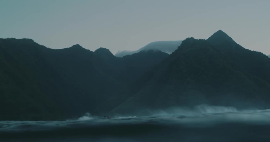 Two people with their surfboards in the open sea with mountainous coastline covered in forest in the background on a cloudy day