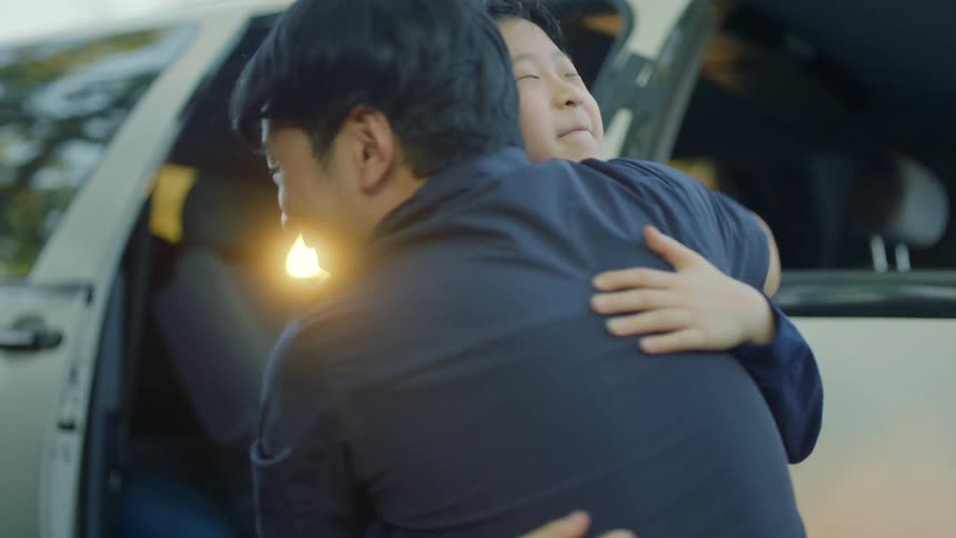 Asian boy runs to adult male and hugs in front of vehicle