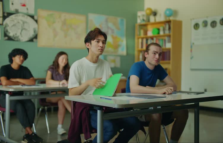 Teenagers in a high school classroom when two students enter one of them sits at a table and the student next to them gives a mocking laugh while the first student raises their hand and responds