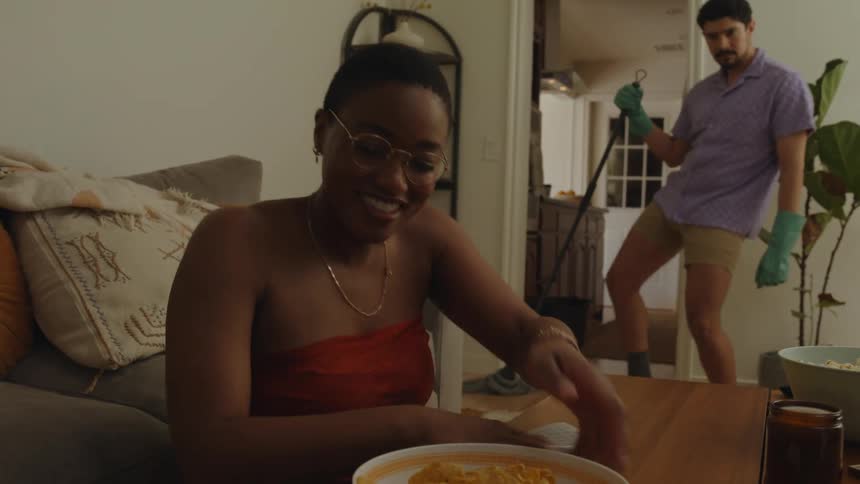 A man and a woman are house cleaning but while she dusts a coffee table he dances while sweeping the floor to make her laugh