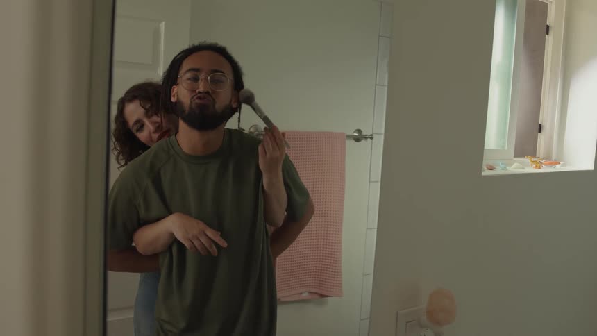 Man and woman stand in bathroom woman uses makeup brush on mans face