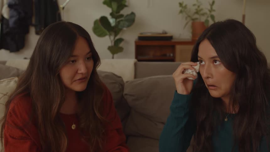 Two teenage girls are sitting together on the couch crying and wiping their eyes with kleenex