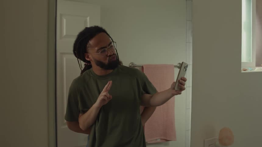 A man is standing in his bathroom holding his phone and smiling as he waves and holds up a peace sign