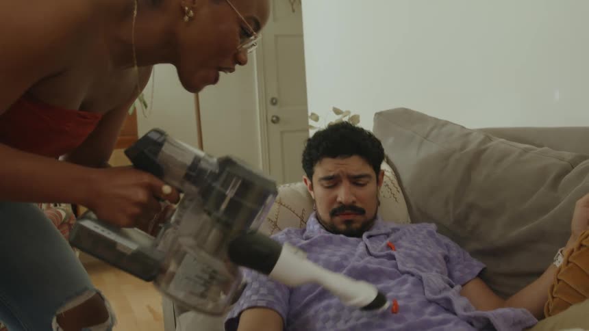 Woman playfully cleans snacks from the chest of a man sleeping in the couch