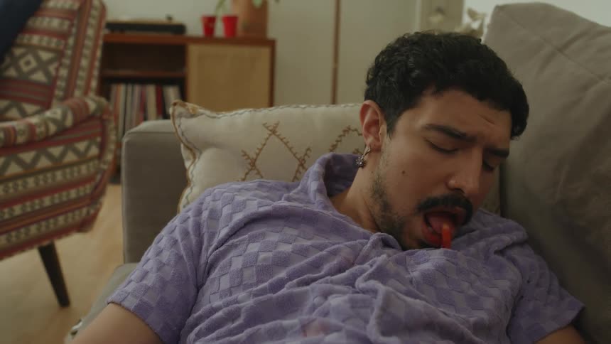 Man lying on the couch while using a vacuum cleaner to suck up a snack that was on his clothes then he wakes up and uses his tongue to eat another snack that was on his clothes