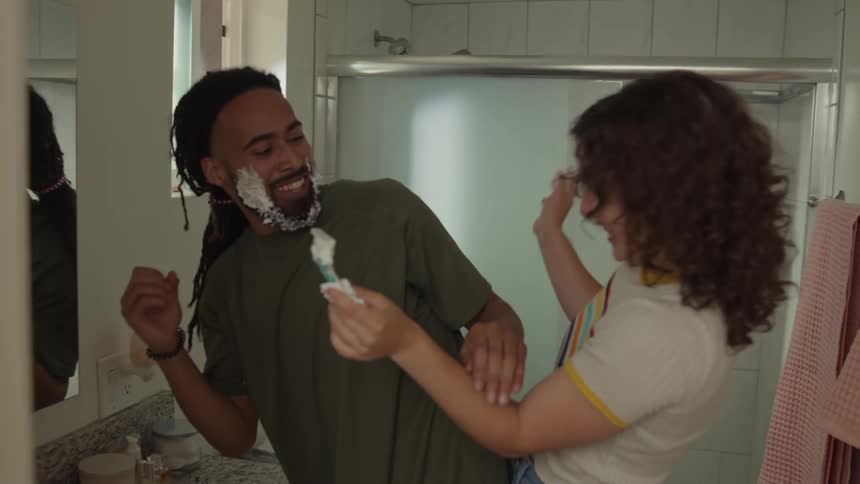 Man looks in bathroom mirror while woman laughs applying shaving cream to his face