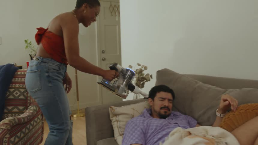 Woman vacuuming her sleeping friend on the couch who wakes up and she continues to vacuum the furniture