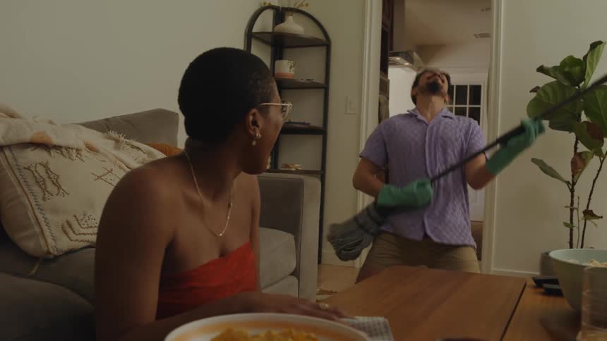 Two people cleaning the interior of a house one of them holding a broom and pretending to play a guitar both of them smiling
