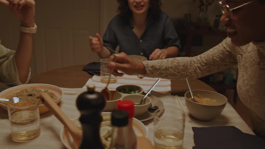 Friends sitting at a table talking and smiling with one of them adding sauce to their bowl of pasta