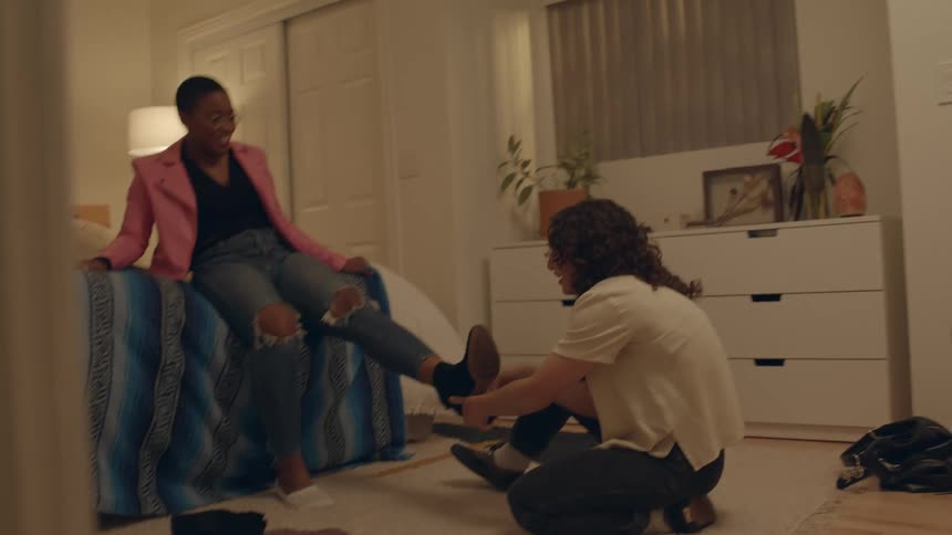 Black woman sitting on a bed laughing while a teenager boy sitting on the floor tries to remove a shoe that got stuck on her foot