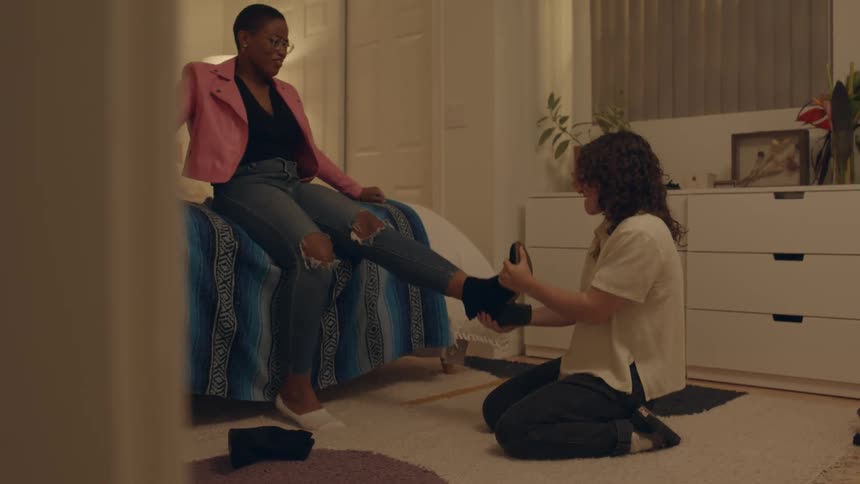 A woman helps her roommate remove a pair of tight boots