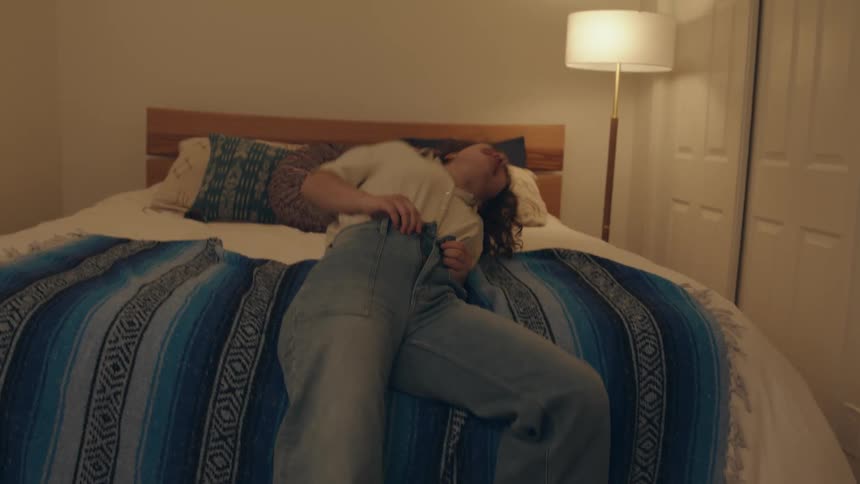 Two teenage girls laugh as one of them struggles to put on a pair of pants and falls onto a bed