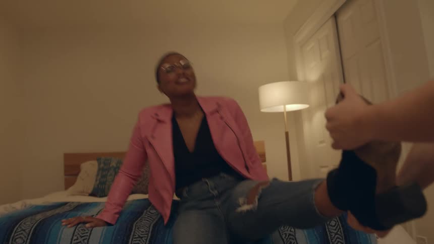 A couple laughs as a man tries to pull a womans boot off while she struggles to stay on the bed
