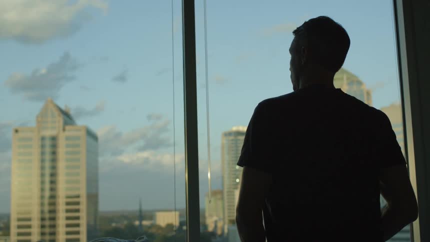 A man is sitting in bed looking at a view of a city out of a window