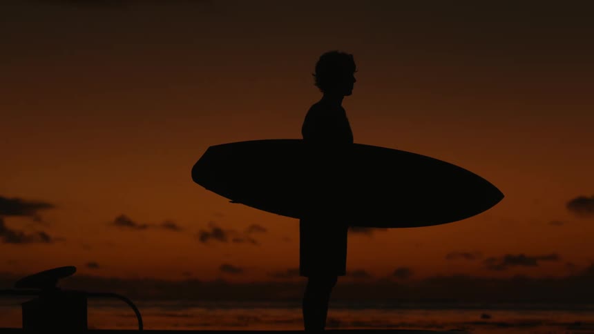 Silhouette of a man holding a surfboard next to the ocean at sunset