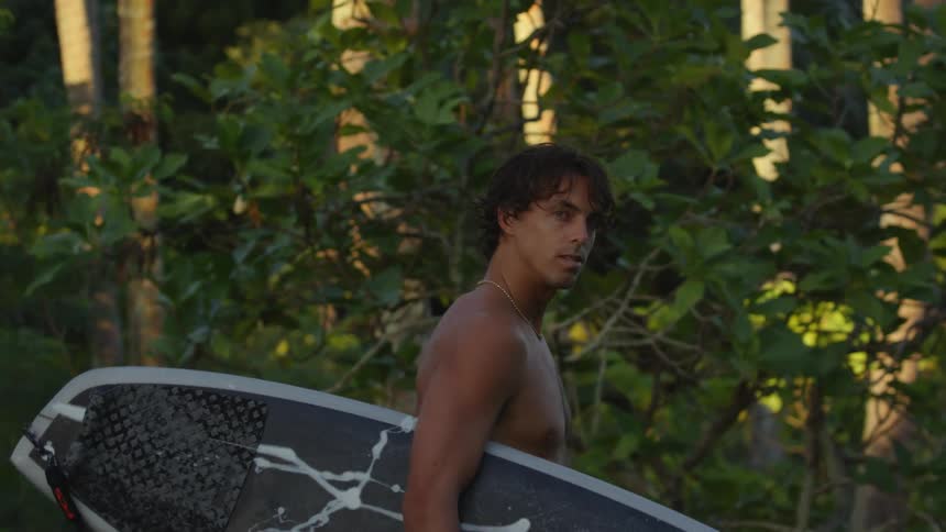 A man is carrying a surf board while walking in a small bush of plant with trees around