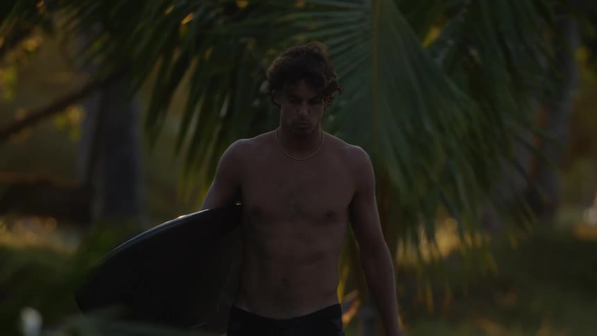 A young man is walking in a tropical area while carrying a surfboard