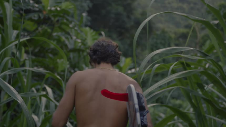 A shirtless man walking alone inside the bush while carrying a surfboard