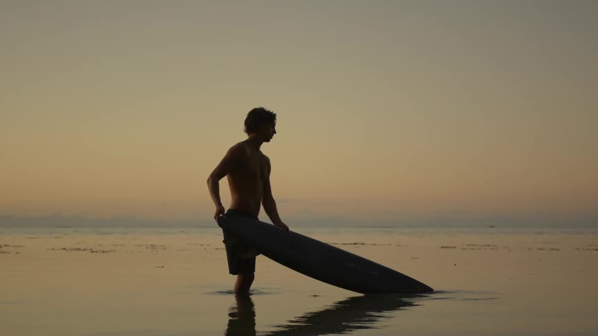 A boy is holding a surf board while standing on the edge of the calm ocean water with no wave in sight