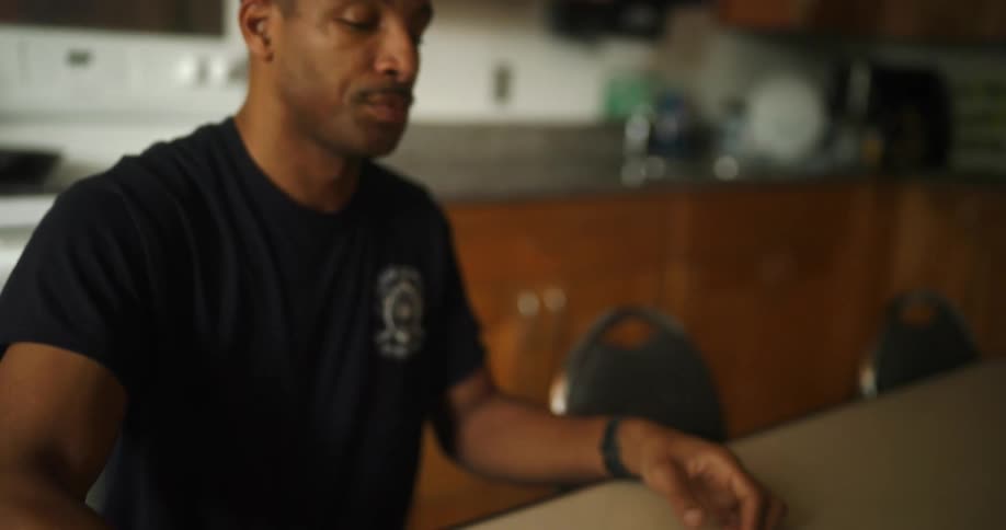 A man is sitting at his kitchen table drinking a cup of coffee and he reaches for his phone on the table