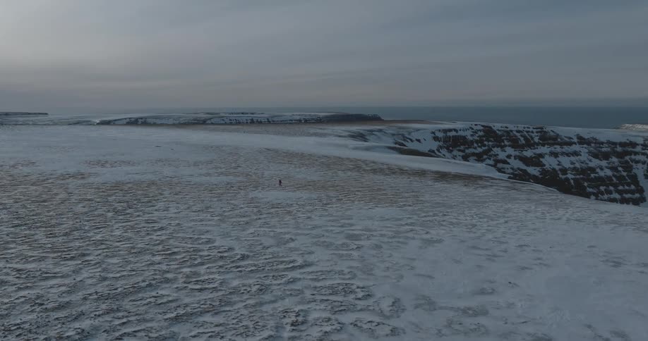 An aerial of snow covered land as a person walks over the terrain near cliff sides