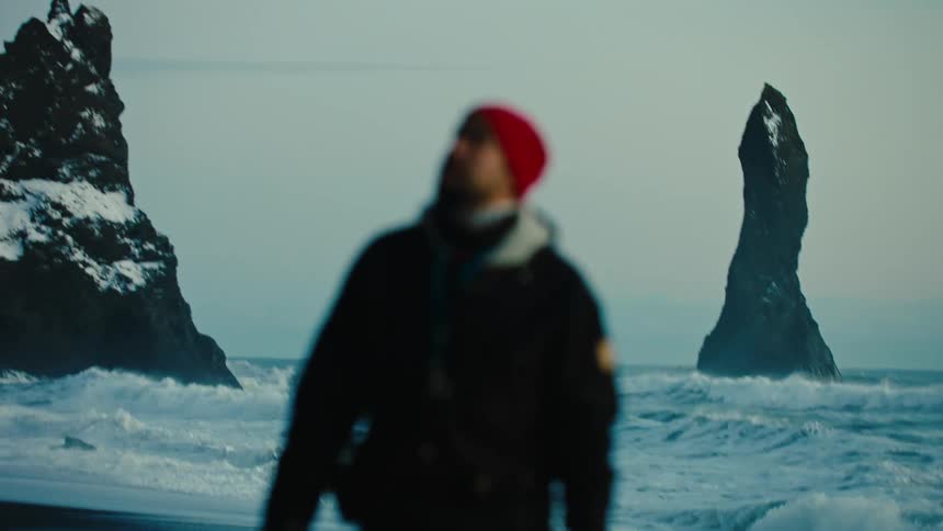A man is walking along the coast as waves roll into shore among snow covered rock formations