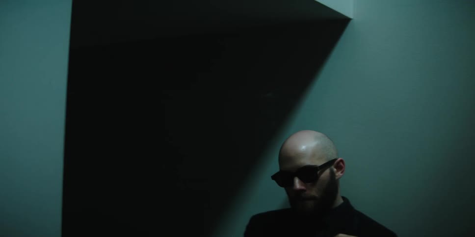 A man sitting against a wall wearing sunglasses while smoking a cigarette