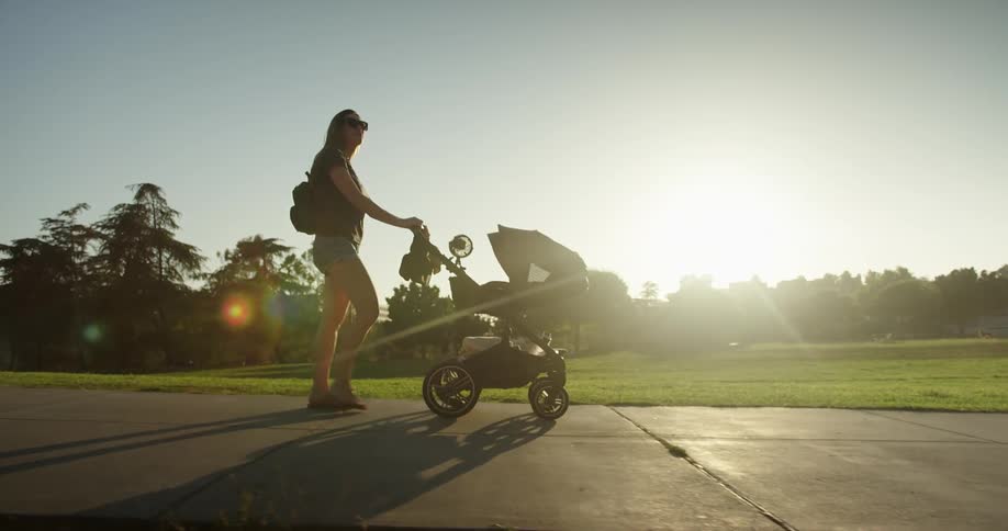 A woman walking a stroller down a sidewalk near an open grass field as the sun rises in the morning