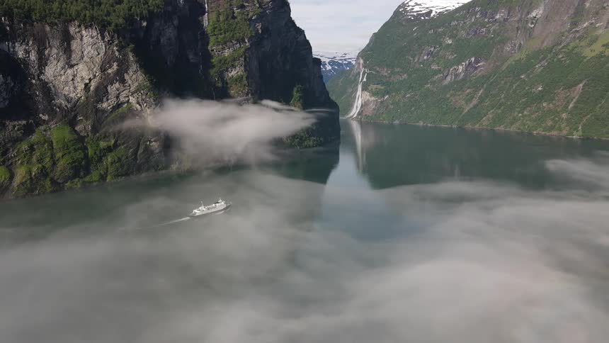 Aerial of a passenger boat sailing through waters in between mountains as fog settles over the surface