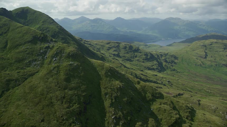 An aerial of a green mountain range surrounding a mountain lake under cloudy skies