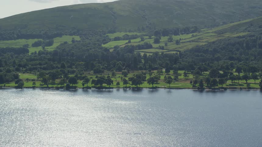 Aerial of a lake along a forest covered hillside as the sun reflects off the water surface