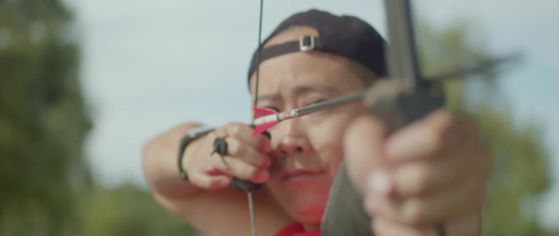 A man wearing a baseball cap holds a bow and aims an arrow