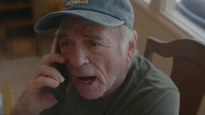 An elderly man in a baseball cap is sitting and talking on a cell phone