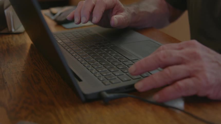An elderly man sitting at a table begins to type on his laptop