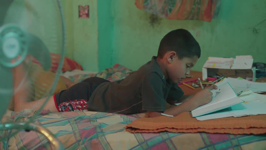 A small boy lying down is drawing in a notebook with a pencil