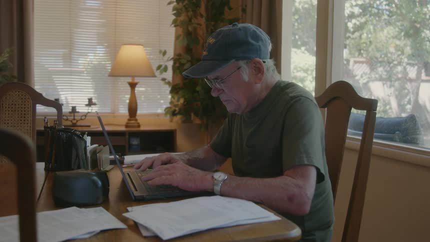 An elder man typing in a laptop while reading some paper
