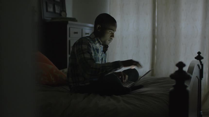 Man types on a laptop while sitting on a bed