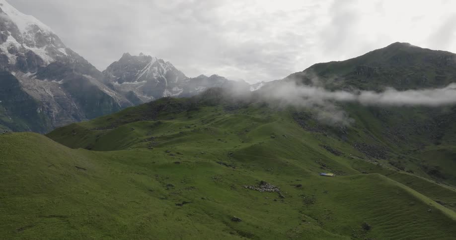 A mountain of tall grass with clouds nearby and behind it mountains with a peak full of snow