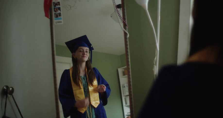 Teenage girl models graduation cap and gown in front of a mirror