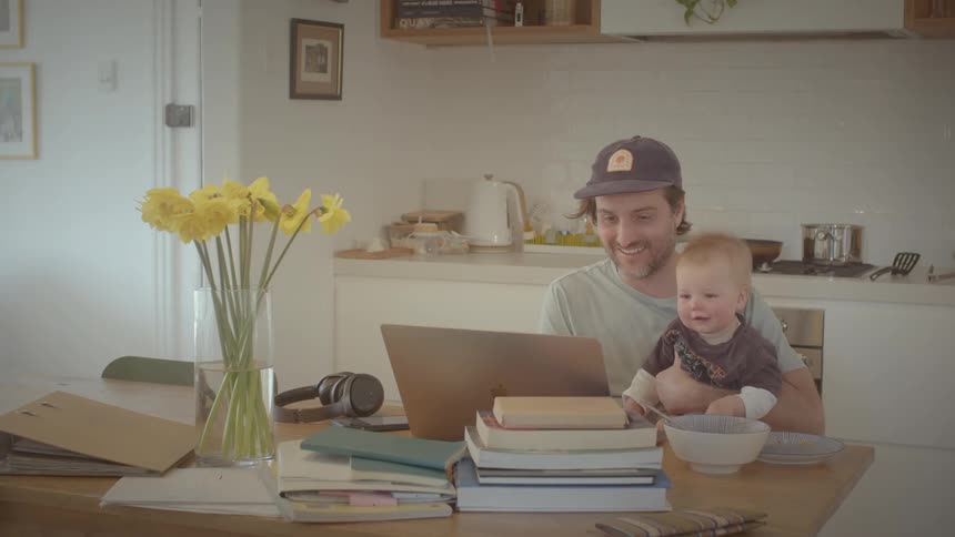 Father and child sit inside the kitchen looking at the screen of a laptop